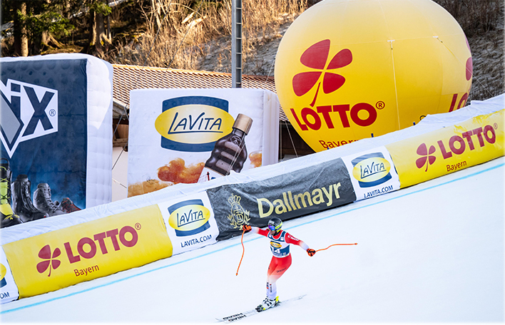 Skirennläufer in rot-weiß-blauer Ausrüstung auf einer Skipiste mit Werbebanden von Lotto Bayern, LaVita und Dallmayr